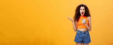 Portrait of displeased pissed and irritated caucasian female. with curly hairstyle in red lipstick and orange cropped top holding smartphone and gesturing with palm annoyed complaining on dumb message