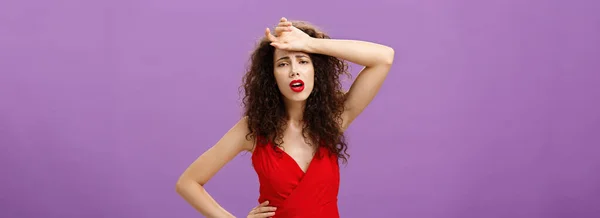 Woman fainting feeling bad whiping sweat of forehead standing drained and exhausted over purple background in red stylish dress expressing gloomy and unhappy feelings wanting some help. Copy space