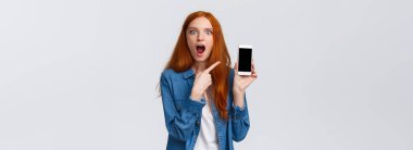 Şok olmuş genç bir kadın eski sevgilisini internetteki sosyal medyada başkasıyla görmüş. Şaşırmış ve etkilenmiş kızıl saçlı kadın akıllı telefon ekranını işaret ediyor, çenesini indir..