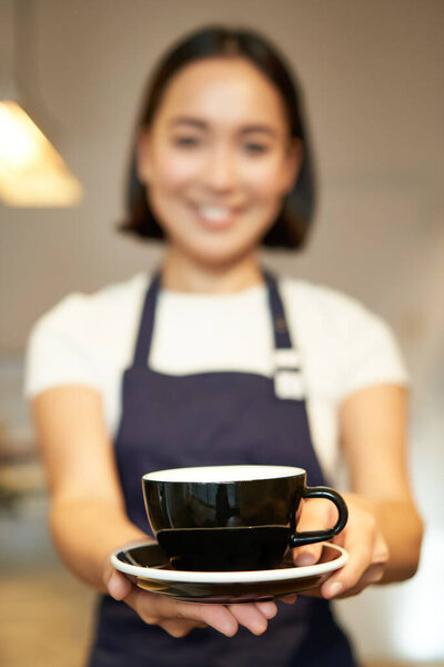 Portrait of smiling asian girl barista, waitress in cafe uniform, giving you cup of coffee, prepare drink for client, looking friendly.