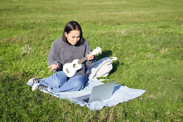 Cute asian girl, musician studying, looks at tutorial on her laptop, online tutor shows how to play ukulele, student sits on grass in park on sunny day.