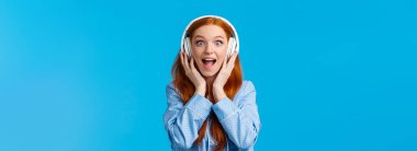 Heyecanlı ve eğlenceli beyaz saçlı, pijama giymiş çekici bir kadın, hayranlık ve şaşkınlıktan bağırıyor, yeni bir şarkı duyuyor, kulaklık takıyor, müzikten hoşlanıyor, mavi arka planda duruyor..