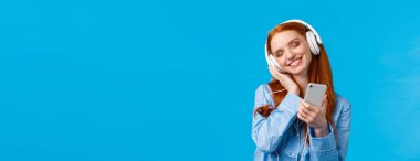 Şefkat, kadınlar ve yaşam tarzı konsepti. Pijama giymiş neşeli kızıl saçlı kadın, başını eğmiş müzik dinlemekten zevk alıyor, akıllı telefon tutuyor ve kulaklık gibi gülümsüyor..