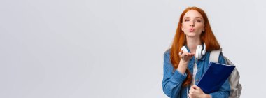 Sevimli ve hassas, şehvetli bir üniversiteli kız, kızıl saçlı şımarık bir öğrenci, hava öpücüğü gönder, kameraya tutkuyla üfle, sırt çantası ve klasörü tut..