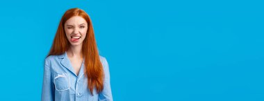 Şımarık ve heyecanlı, gece elbisesi içinde güzel saçlı bir kadın, neşeyle göz kırpan ve gülümseyen bir kamera, harika hissediyor, pozitif duygularını ifade ediyor, mavi arka planda duruyor..