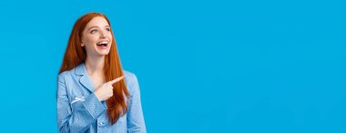 Gecelik giymiş, pijamayla uyuyan, üst sol köşeyi işaret eden, mutlu bir şekilde gülen, mavi arka planda duran, beyaz saçlı, hoş ve eğlenceli bir kız..