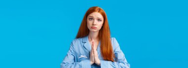 Lütfen, sana yalvarıyorum. Aptal ve tedirgin kızıl saçlı beyaz kız, yardım istiyor, af diliyor ya da başka bir şey istiyor, eğik kafalı, üzgün suratlı, mavi arka planda duruyor..