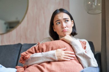 Kadınlar ve refah kavramı. Hüzünlü ve kasvetli Asyalı bir kadının portresi mutsuz bir şey düşünüyor, kanepede yastığa sarılıyor ve sıkıntısını dile getiriyor..