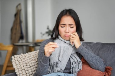 Kız termometreye kızgın görünüyor, ateşi var, evde hasta yatıyor, kendini iyi hissetmiyor, atkıda oturuyor..