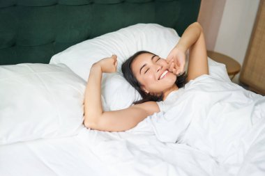 Mutlu sabahlar. Güzel Asyalı bir kız, beyaz bir yatakta kollarını açıp mutlu bir şekilde gülümseyen bir otel elbisesiyle uyanır..