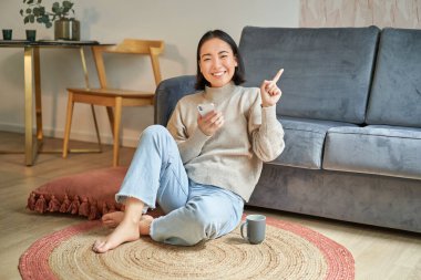 Modern Koreli genç kadın portresi yerde oturuyor, fotokopi alanını işaret ediyor, afişi veya reklamı gösteriyor, akıllı telefonu uygulama olarak tutuyor.
