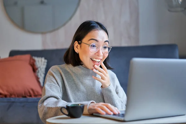 Portrait of asian woman in glasses, sitting with laptop and looking surprised, amazed by promotion on computer.