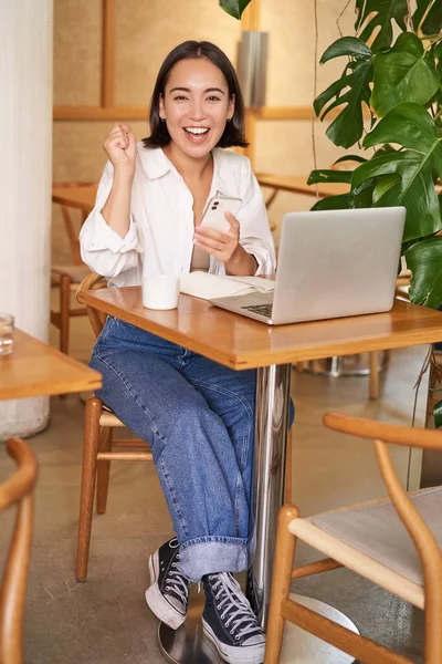 Full body shot of young asian girl sits in cafe with laptop, sees good news on smartphone and celebrates, triumphs from goal or achievement.