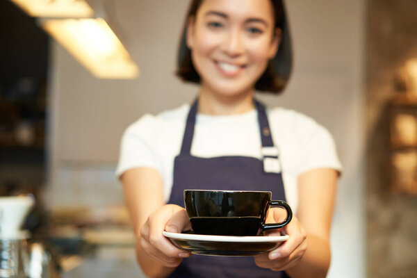 Smiling asian girl barista, standing in apron uniform, giving you cup of coffee, made a drink for client in cafe.