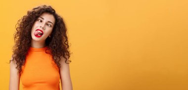 Kız kaygısız geziyor. Sıkıcı toplantıda kendini eğlendirmeye çalışmaktan sıkılmış. Dilini çıkarmış, gözlerini kısmış, başını eğmiş turuncu arka planda çocukça ve çocukça poz vermiş..