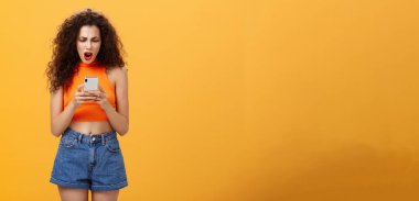 Sosyal ağ profilindeki düşük fotoğraf oranından memnun olmayan ve gücenen bir kadın, hayal kırıklığı yüzünden soluk soluğa kalıyor ve turuncu arka planda duran akıllı telefon ekranından memnun kalmamış görünüyor.