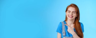 Neşeli, kaygısız, kızıl saçlı, samimi bir gülümseme, solu işaret eden, fotokopi alanını tanıtan, neşe dolu, kapalı gözler, beyaz dişlek bir gülümseme, neşeli, neşeli, pozitif bir ruh hali, mavi arka plan..