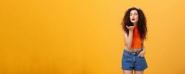 Şık, cilveli, kendine güvenen, kıvırcık saçlı, kırmızı rujlu, kameraya karşı şehvetli bir şekilde gülümseyen bir kadın.