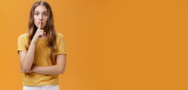 Oyun sırasında sessiz ol. Ciddi ve kararlı, dalgalı saçlı, sessiz jest yapan, ağzı işaret parmaklı, sessiz kalan, turuncu duvarda sır saklayan genç bir kadın..