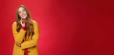 İyi görünümlü kaygısız ve mutlu, gevşemiş, sarı renkli, şık ceketli, yüzü ve başını eğmiş kızıl saçlı kız arkadaş olumlu duygularla gülümsüyor, kırmızı arka plana karşı neşeli poz veriyor..