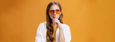 Uzun, dalgalı, güneş gözlüklü, bluzlu, sırıtan, sol üst köşede kararlı bakışlı, çenesini ovuşturup turuncu duvarın üzerinde poz vermeyi düşünen düşünceli, şık bir parti kızı..