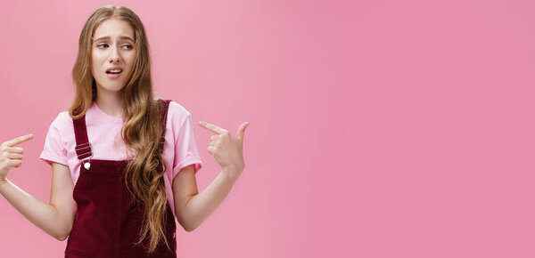 Self-assured arrogant young female student with high ego standing cocky and brag about herself pointing at her with cool snobbish look looking away to right with contempt posing over pink wall. Body