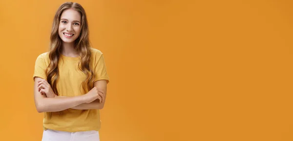 Lifestyle. Portrait of kind and friendly charming young female student in casual t-shirt with wavy natural long hair holding hands crossed on chest smiling broadly and carefree at camera over orange