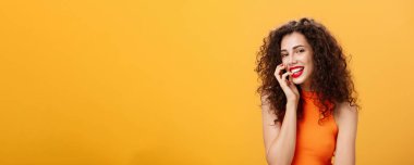 Çekici, aptal ve kaygısız bir kadının belden yukarısına vuruşu. Kıvırcık saçlı, üst parmağıyla ısıran, şehvetli bir şekilde gülümseyen ve kameraya cilveli seksi ve turuncu arka planda gözüken bir kadın.