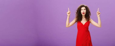 Kafası karışmış Avrupalı çekici bir kadın. Kıvırcık saçlı, kırmızı elbiseli, ağzı açık, kollarını kaldırmış düşünceli ve meraklı bakışları olan, mor arka planda sıkıntılı biri..