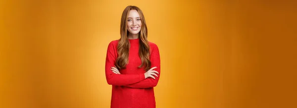 Portrait of elegant and friendly, polite redhead female in red knitted dress holding hands crossed over body in confident and relaxed pose smiling satisfied offering help over orange background.