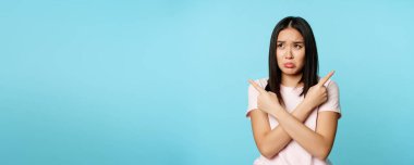 Kafası karışmış ve üzgün Asyalı kız yan bakıyor, cahil ya da kararsız görünüyor, seçim yapmakta zorlanıyor, asık suratını asıyor, mavi arka plan.