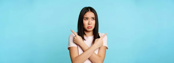 Confused and sad asian girl pointing sideways, looking clueless or indecisive, problem with choosing, pulling long upset face, blue background.