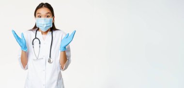 Mutlu Asyalı kadın doktor el çırpıyor, şaşırtıcı haberlere hevesli bakıyor, tıbbi maske ve lastik eldiven takıyor, beyaz arka plan.