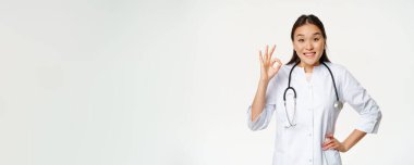 Gülümseyen Asyalı doktor, sağlık önlüğü giyiyor. Üniformalı kadın hastane çalışanı, beyaz arka planda durarak smth öneriyor..