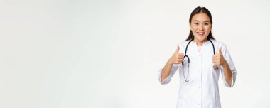 Gülümseyen Asyalı kadın doktor, tıbbi bornoz giyiyor, başparmaklarını kaldırıyor, sağlık ürünleri öneriyor, beyaz arka planda duruyor..