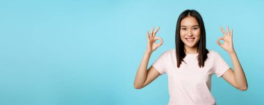 Hiç sorun değil. Gülümseyen Asyalı kadın iyi işaretler gösteriyor, onay jesti, memnun ve memnun, mavi arka planda duruyor..
