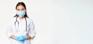 Covid-19 ve sağlık hizmeti konsepti. Tıbbi maskeli genç Asyalı kadın doktor, lastik eldivenler ve klinik üniforması yardım etmeye hazır, dinleyen hasta, beyaz bir geçmişi var..