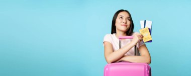 Seyahat hayalleri kuran mutlu Asyalı kadın turist, bavula yaslan, elinde covid-19 uluslararası aşı sertifikası, iki bilet ve pasaport, seyahat düşüncesi, mavi arka plan.