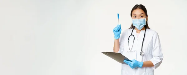 Excited female doctor, asian physician holding clipboard and raising pen up, found solution or idea, standing in medical face mask over white background.