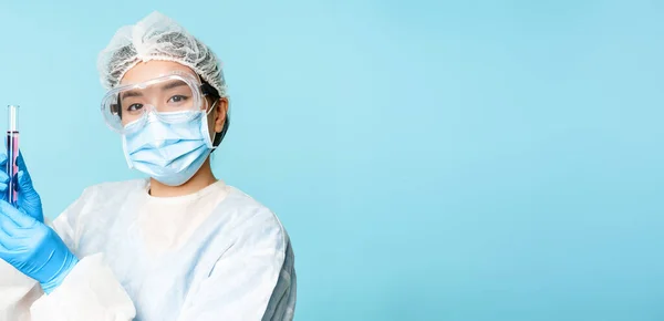 Asian nurse or lab worker in personal protective equipment, showing test sample tube, standing in medical face mask over blue background.