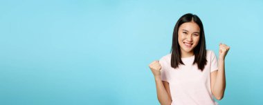 Hevesli, sevimli Asyalı kız kazanıyor, harika haberlerle seviniyor, zafer dansı yapıyor, gülümsüyor, mutlu, mavi arka planda duruyor..