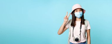 Tıbbi maske takan mutlu Koreli kadın, fotoğraf makinesi, barış işaretleri gösteren, covid-19 salgını sırasında seyahat eden, yurt dışında tatil yapan, mavi arka plan.