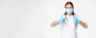 Sağlık ve Coperid-19 önleyici önlemler kavramı. Yüz maskesi ve lastik eldiven takmış Asyalı bir doktor, baş parmak gösteriyor, beyaz arka planda üniforma giyiyor..
