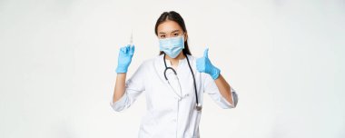 Covid-19 aşısı ve sağlık sigortası. Tıbbi maske ve eldiven takan Asyalı kadın doktor elinde aşı şırıngası ve baş parmağı havada, beyaz arka plan.