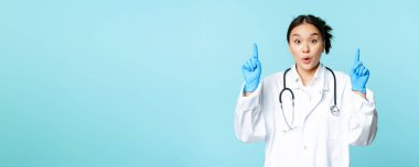 Heyecanlı Asyalı tıp çalışanı, kadın doktor parmaklarını havaya kaldırıyor, steril eldiven ve üniforma giyiyor, mavi arka planda duruyor..
