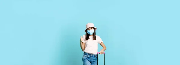 Happy korean woman in medical face mask, thumbs up, posing with cute suitcare, tourist going on vacation, travelling during covid-19 pandemic concept, blue background.