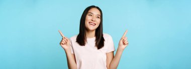 Gülümseyen esmer Asyalı kadın, iki tarafı da gösteriyor ve gülüyor, mağazayı seçiyor, mavi arka planda duruyor..