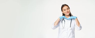 Gülümseyen Asyalı doktor görüntüsü, dermatolog sağlıklı bir cilt ve kameraya sevimli bir poz gösteriyor, beyaz arka planda sağlık üniforması içinde duruyor..