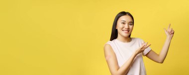 Covid-19 aşı konsepti. Omzunda yama olan gülümseyen Asyalı kız, sağlık afişini işaret ediyor, sarı arka plan.