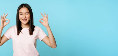 Gülümseyen Koreli kadın göz kırpıyor, iyi işaretler gösteriyor, şirket ya da mağaza tavsiye ediyor, mavi arka planda tişört giyiyor..